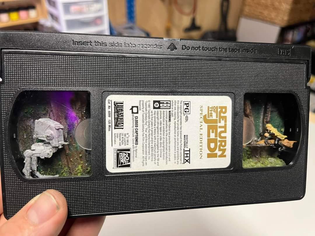 The meme is a photo of a VHS tape with a label that says "Return of the Jedi" and a small window on the front that shows a miniature scene of a Star Wars character (it appears to be a Stormtrooper) in a forest-like setting. The scene is small and detailed, with tiny trees and rocks, and the character is posed as if it's about to take a step into the scene. The caption for the meme is "I finally found the last tape in the attic" which implies that the person has discovered an old, nostalgic item that brings back memories.